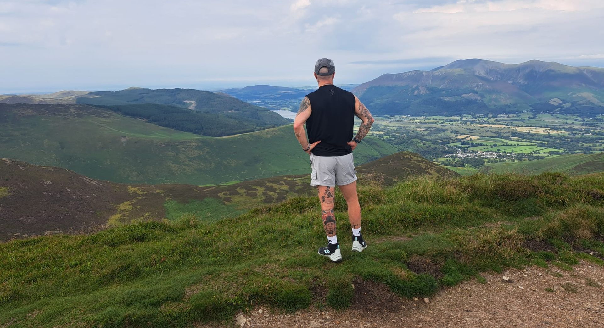 Person standing on a grassy hill overlooking a valley and distant mountains.