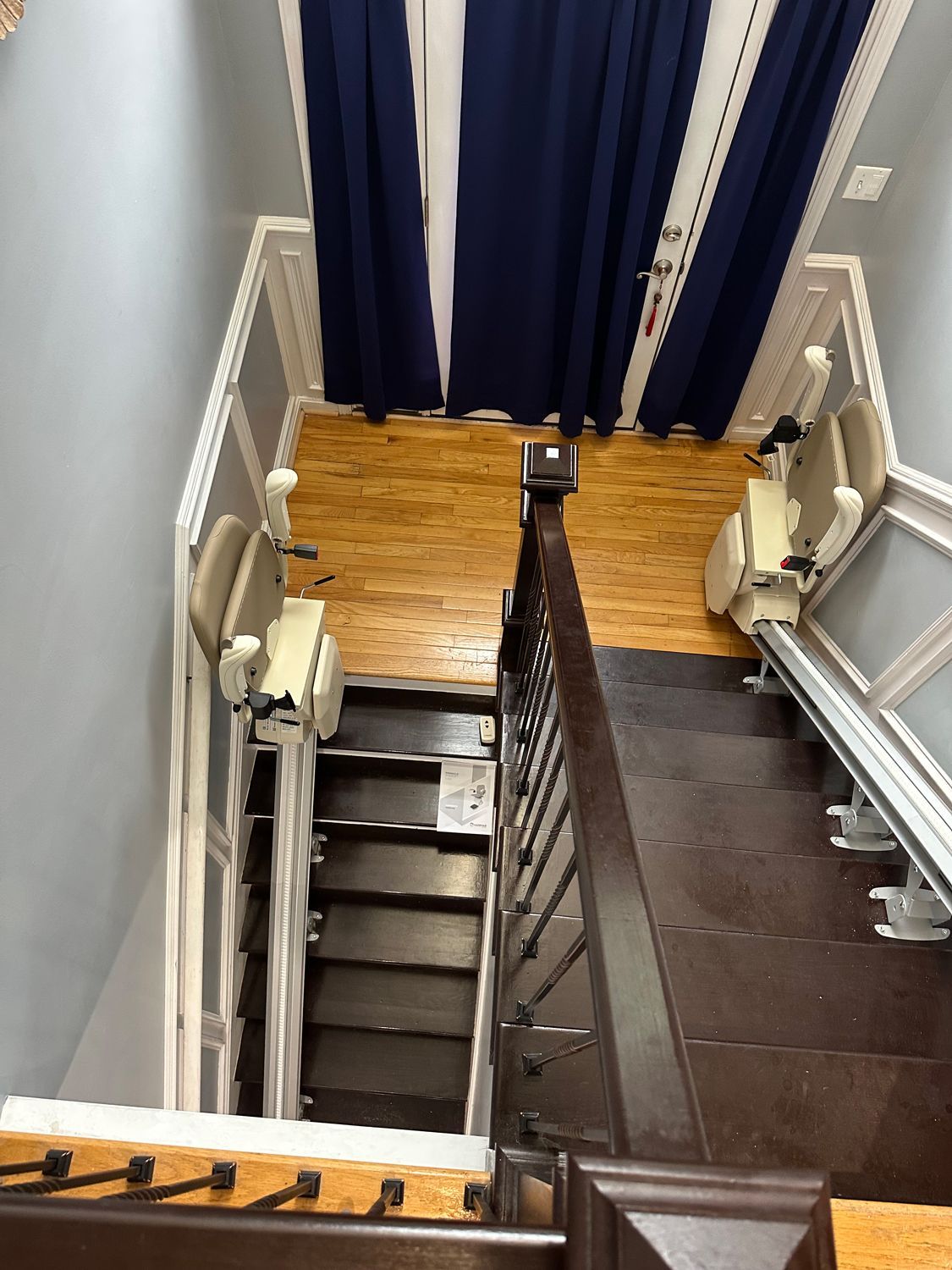 Narrow stairwell with dark steps, white railings, and a small landing with dark curtains and wood flooring.