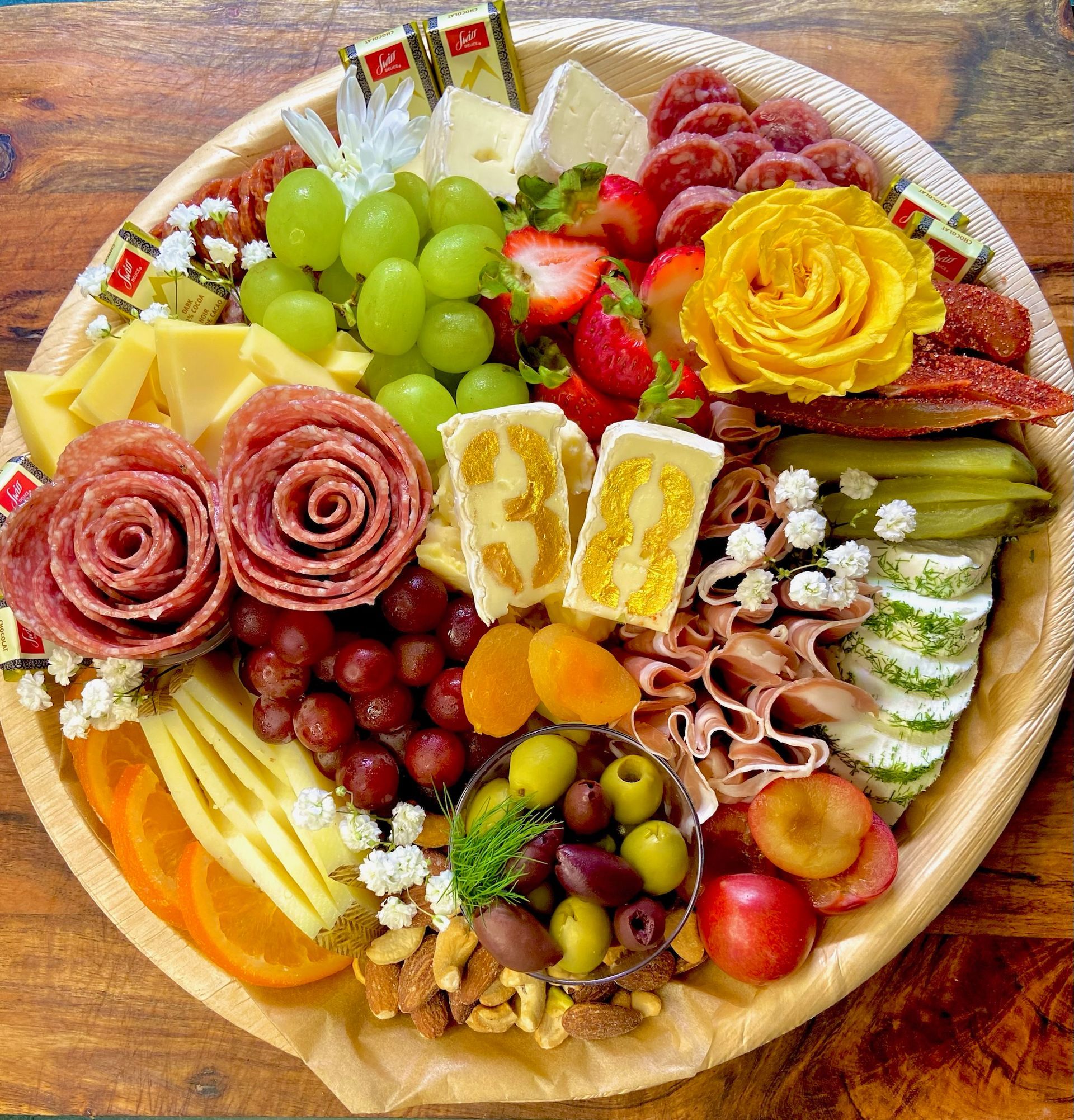 A wooden plate topped with meat , cheese , fruit , nuts and flowers.