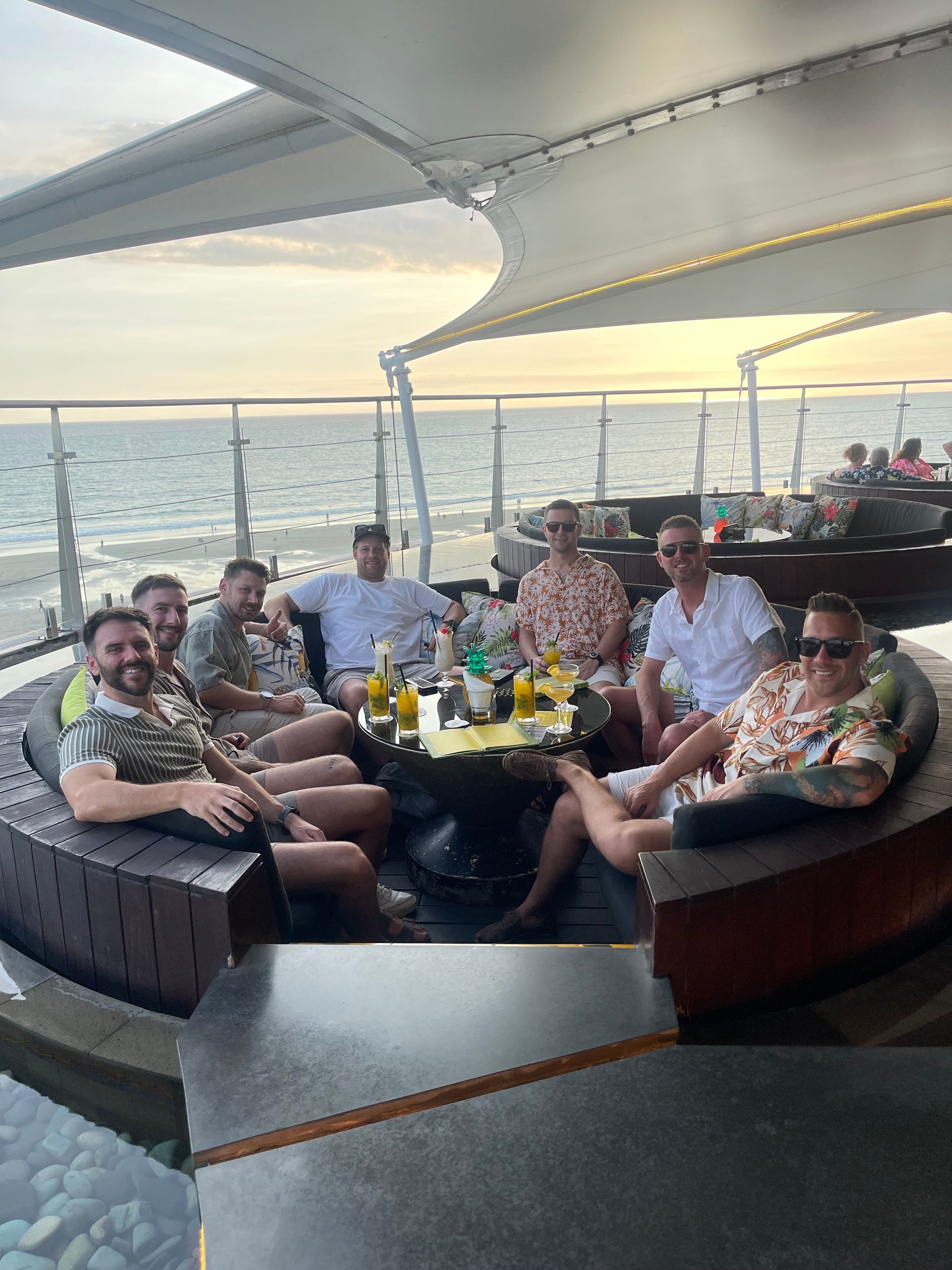 A group of men are sitting around a table on a balcony overlooking the ocean.
