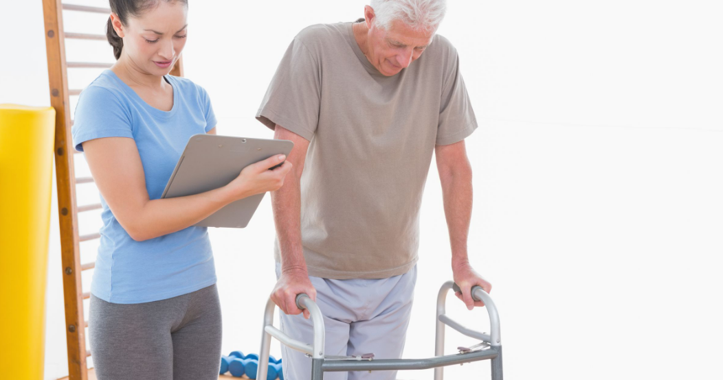 An elderly man is using a walker while a woman looks at a clipboard.