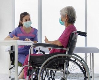 A nurse wearing a mask is talking to an elderly woman in a wheelchair.