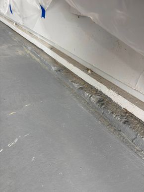 Close-up of a white wall with blue tape and a chipped, dirty floor edge along a gray surface