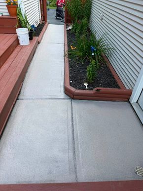 Narrow concrete walkway beside a house with raised planter beds and small garden plants.