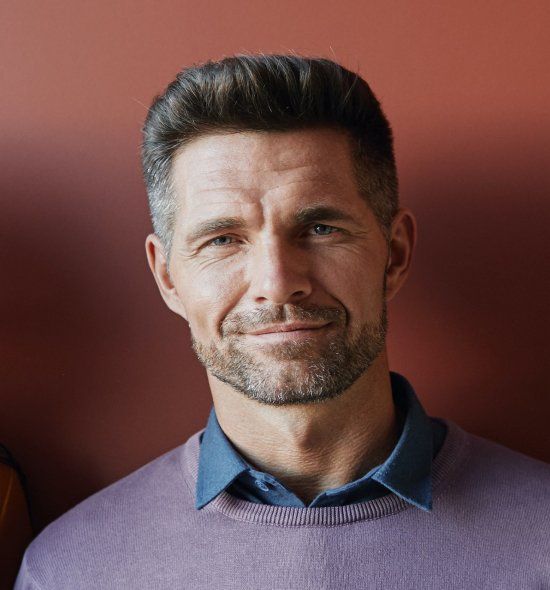 A person with short, dark hair and a light beard, wearing a purple sweater over a blue shirt against a terracotta background.