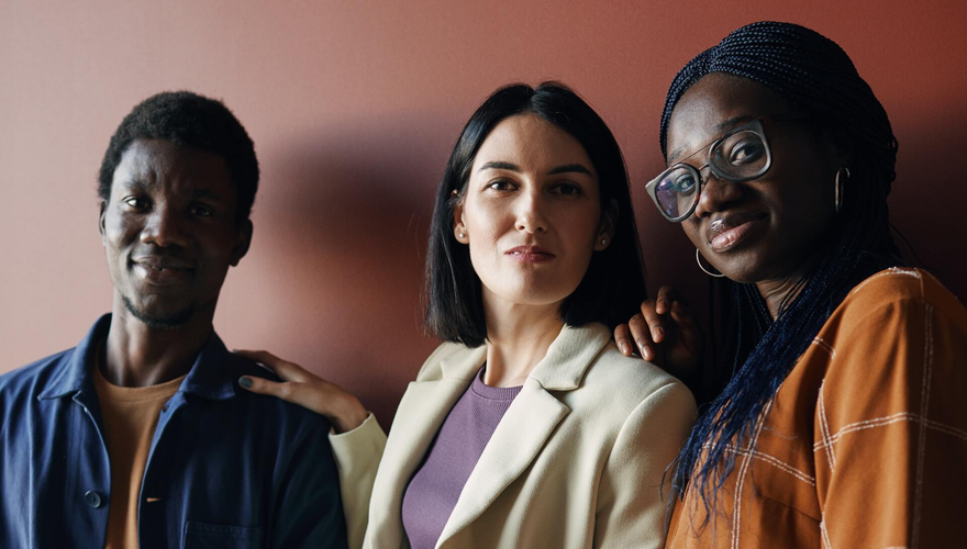 Three diverse people smiling, standing closely together against a solid reddish-brown wall.
