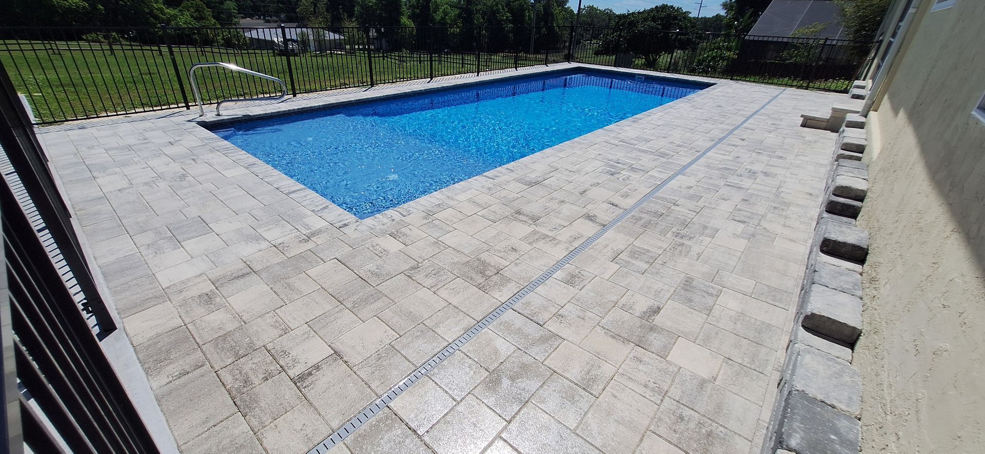 A large swimming pool is surrounded by a patio and a fence.