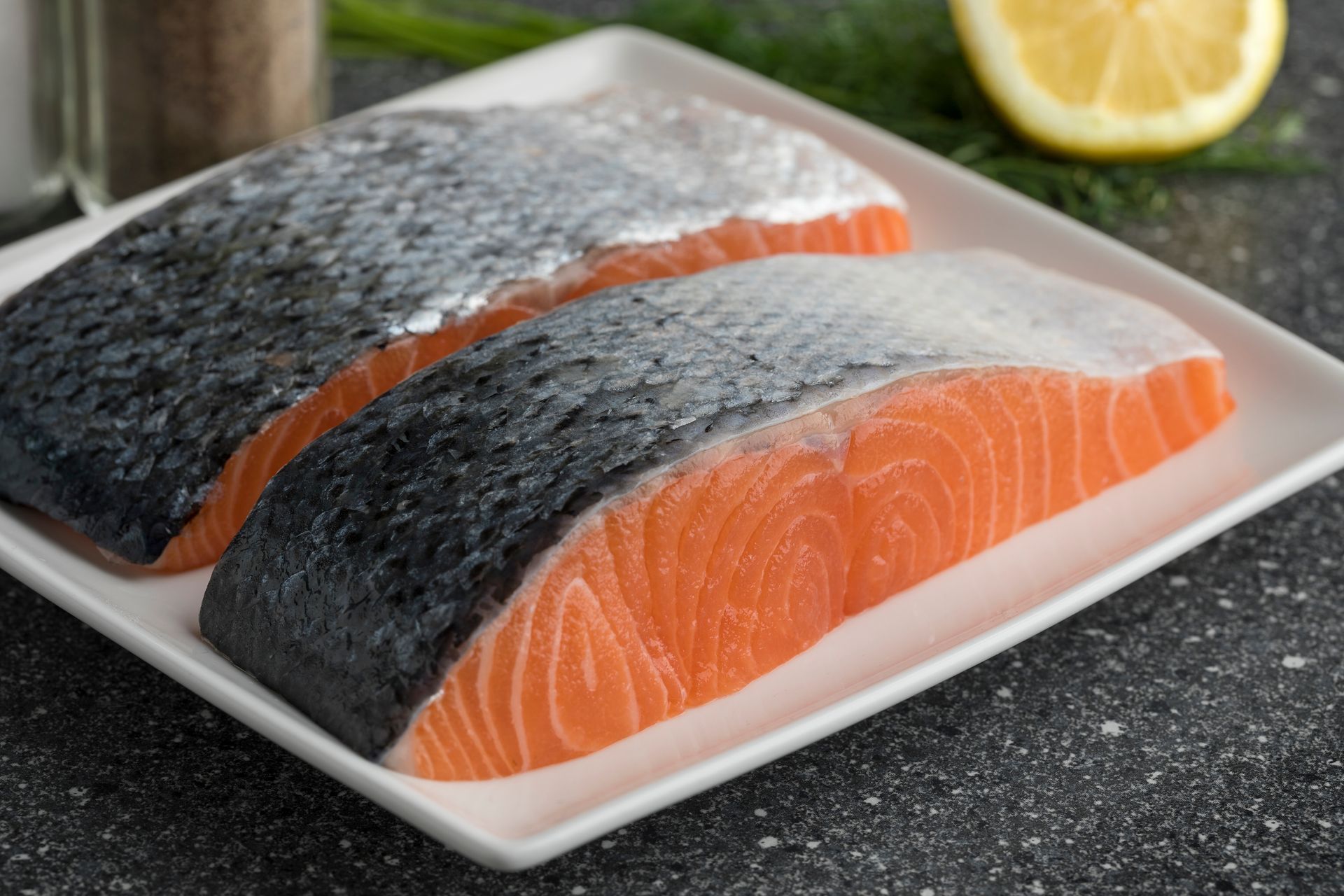Two raw salmon fillets on a white plate with a lemon wedge on a countertop