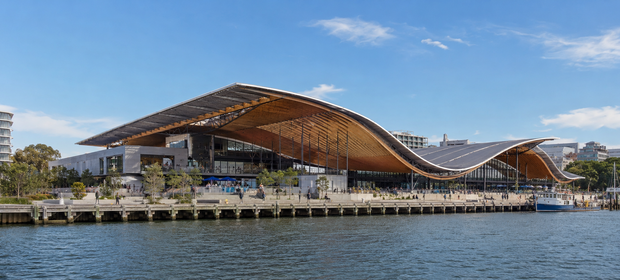 A modern waterfront pavilion with a sweeping wooden roof beside calm water under a clear sky.
Sydney Fish Market.