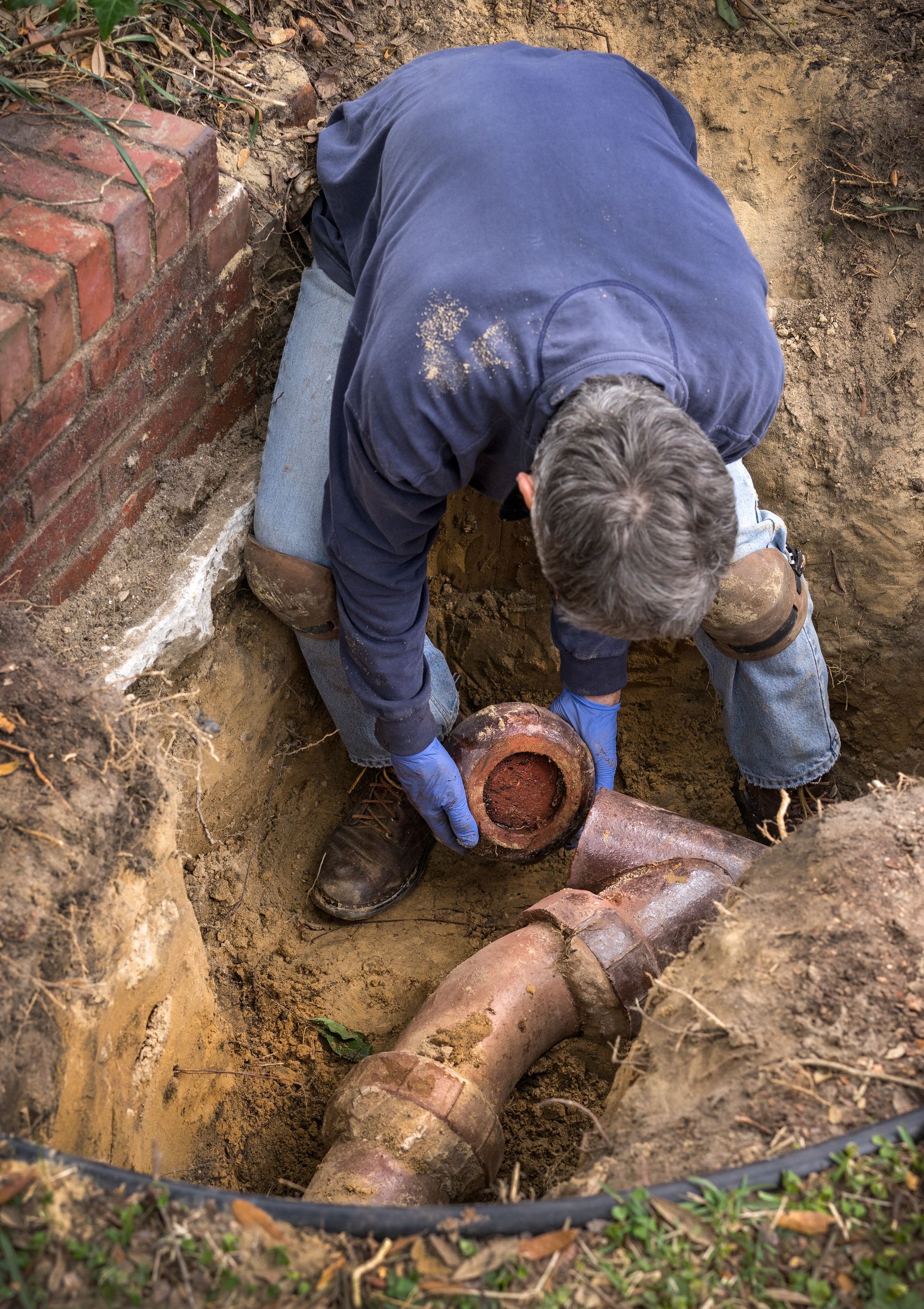 A plumber is working in a dirt trench to replace a corroded clay sewer pipe at a home.