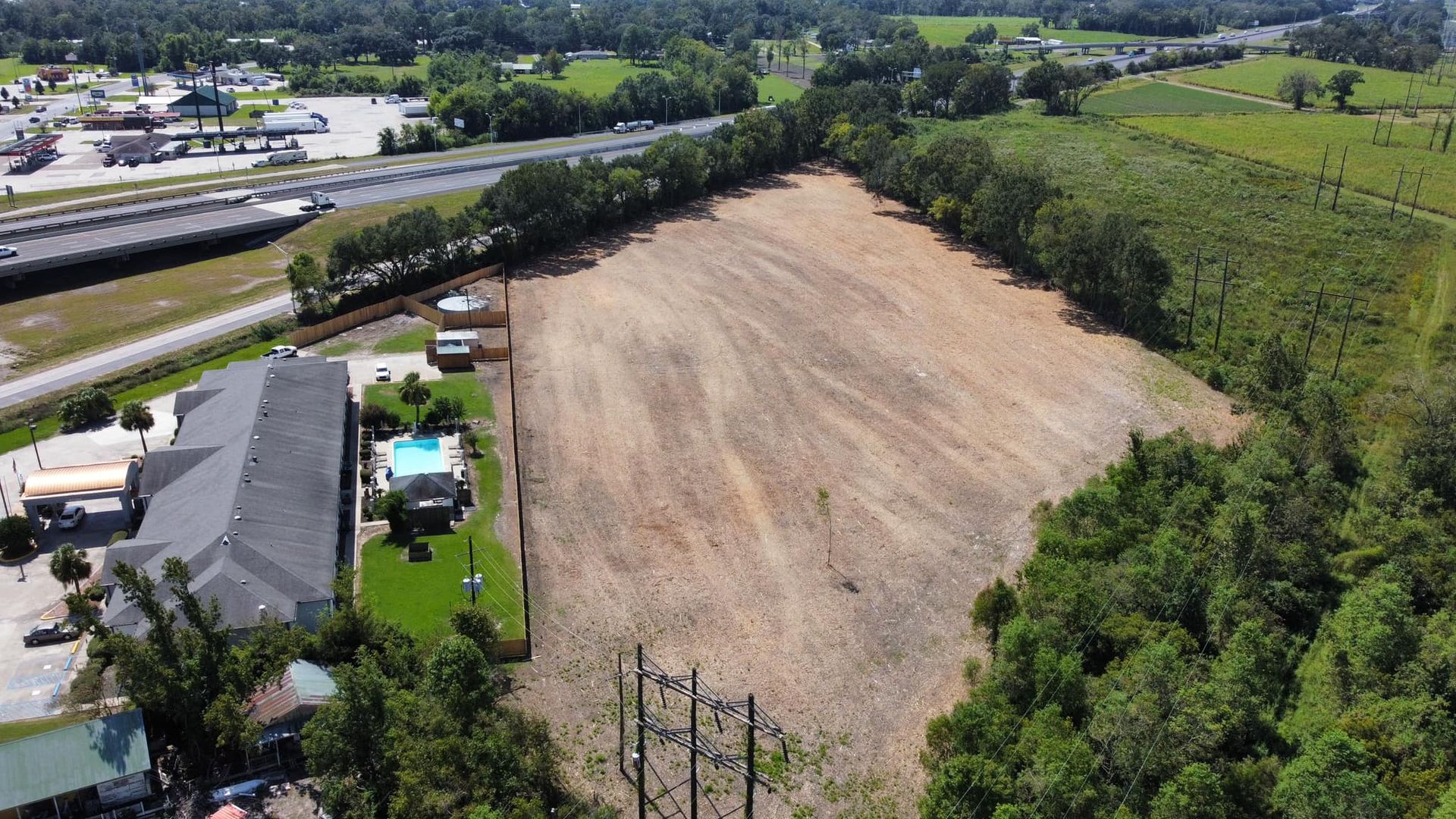 Aerial view of a cleared dirt lot beside houses, a road, and surrounding green trees.