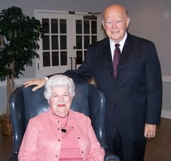 Older couple posing indoors; woman seated in blue chair, man standing beside her in a suit.