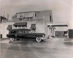 Vintage black car parked outside a retro motel with awnings and a covered driveway.