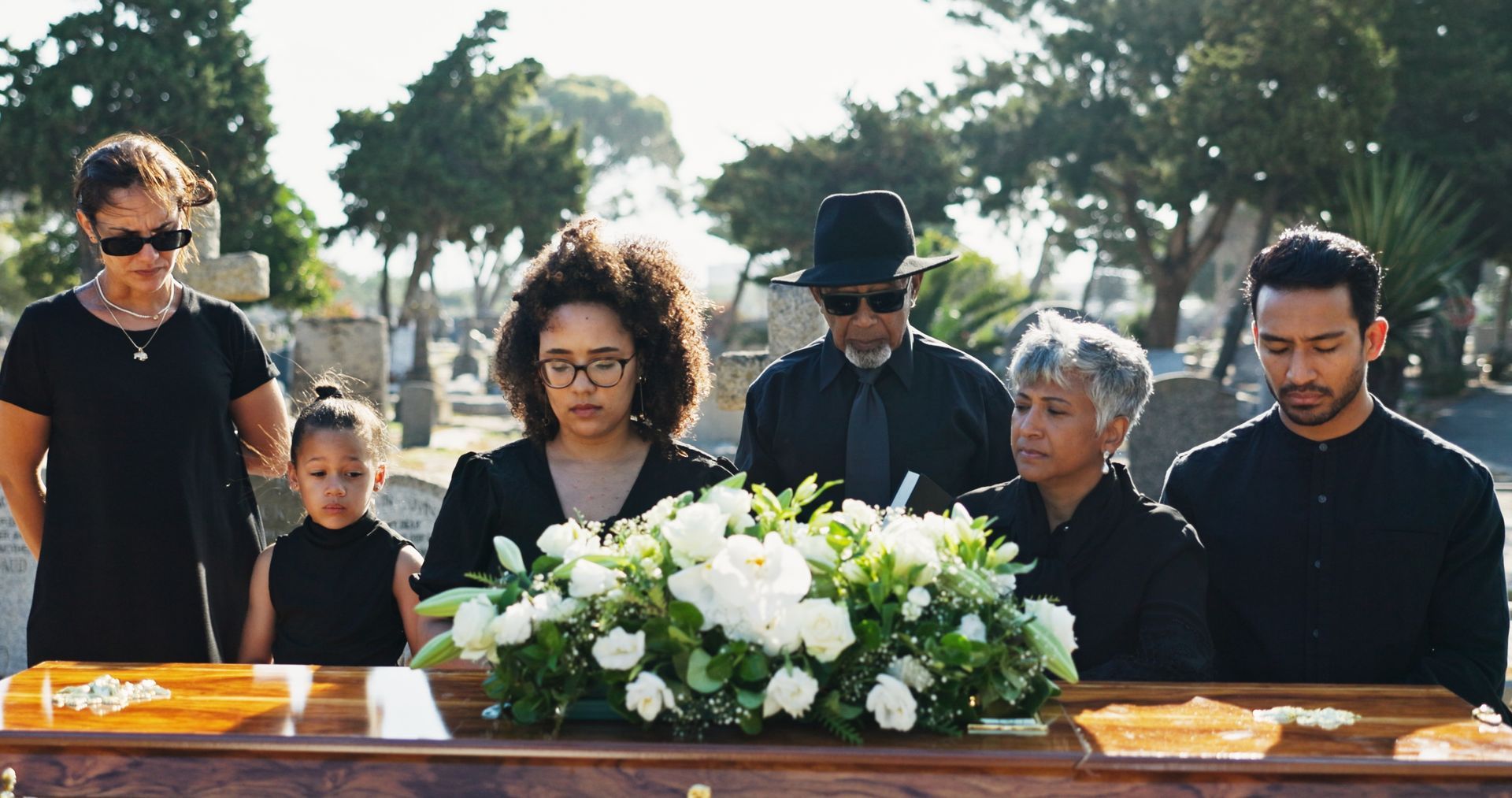 Mourners gather around a casket adorned with white flowers at an outdoor funeral