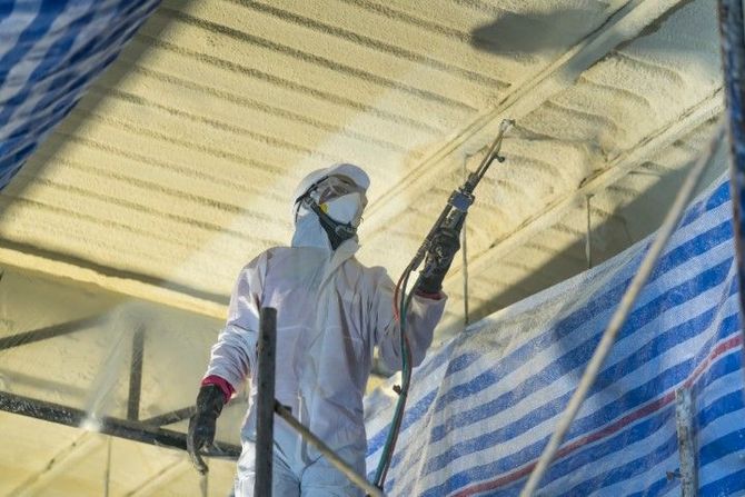 Worker in protective coveralls spraying insulation inside a building, with blue tarps overhead.