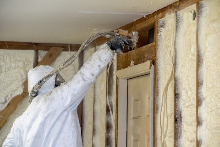 Worker in white protective suit spray-insulating an attic wall with foam.