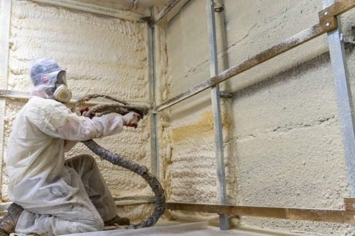 Worker in protective gear spray-foaming insulation on a wall frame inside a room