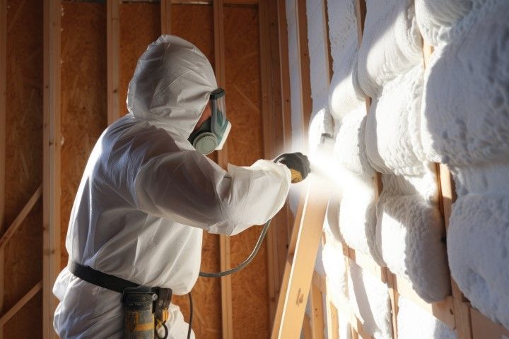 Worker in white protective suit spray-insulating a wall inside a wooden-framed room.