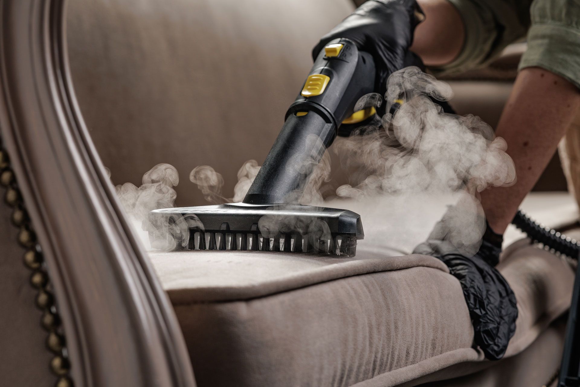 Person steam-cleans a beige upholstered chair with a handheld vacuum