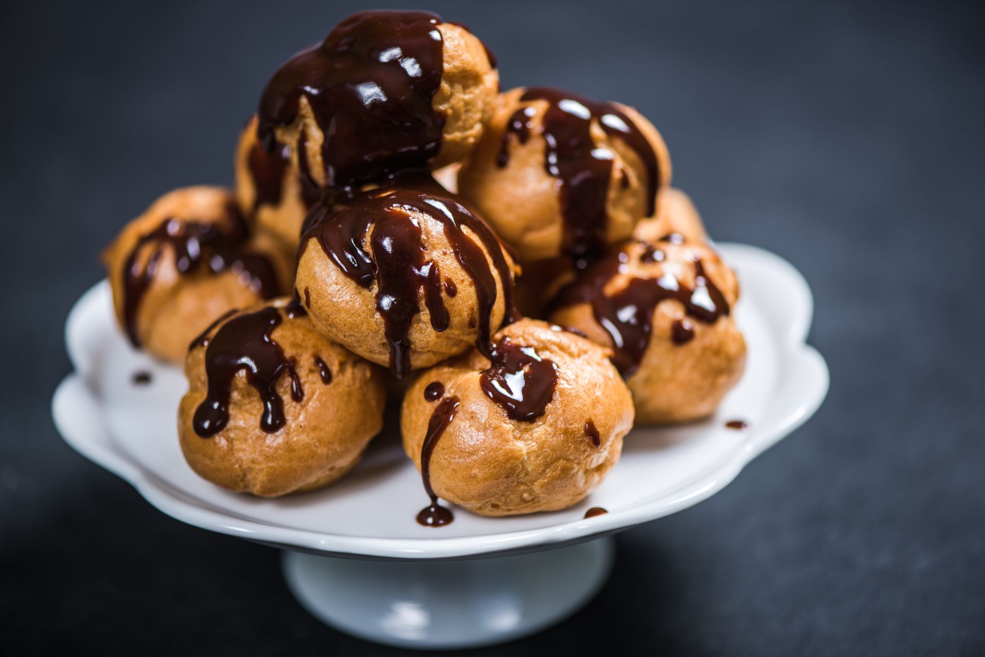 Plate of cream puffs drizzled with chocolate sauce on a white pedestal stand