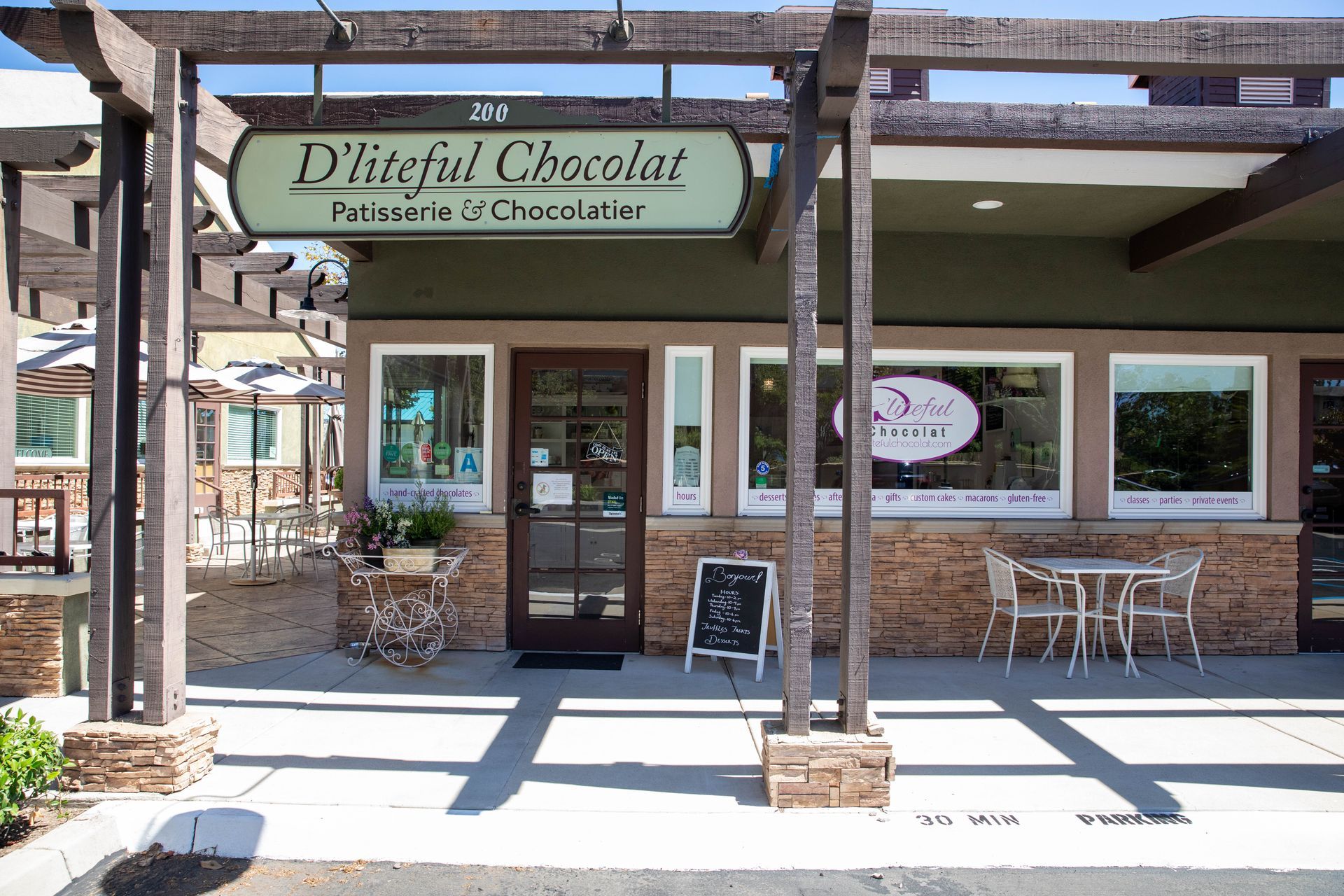 Chocolate shop storefront with green awning, brick facade, and outdoor seating under a wooden pergola.