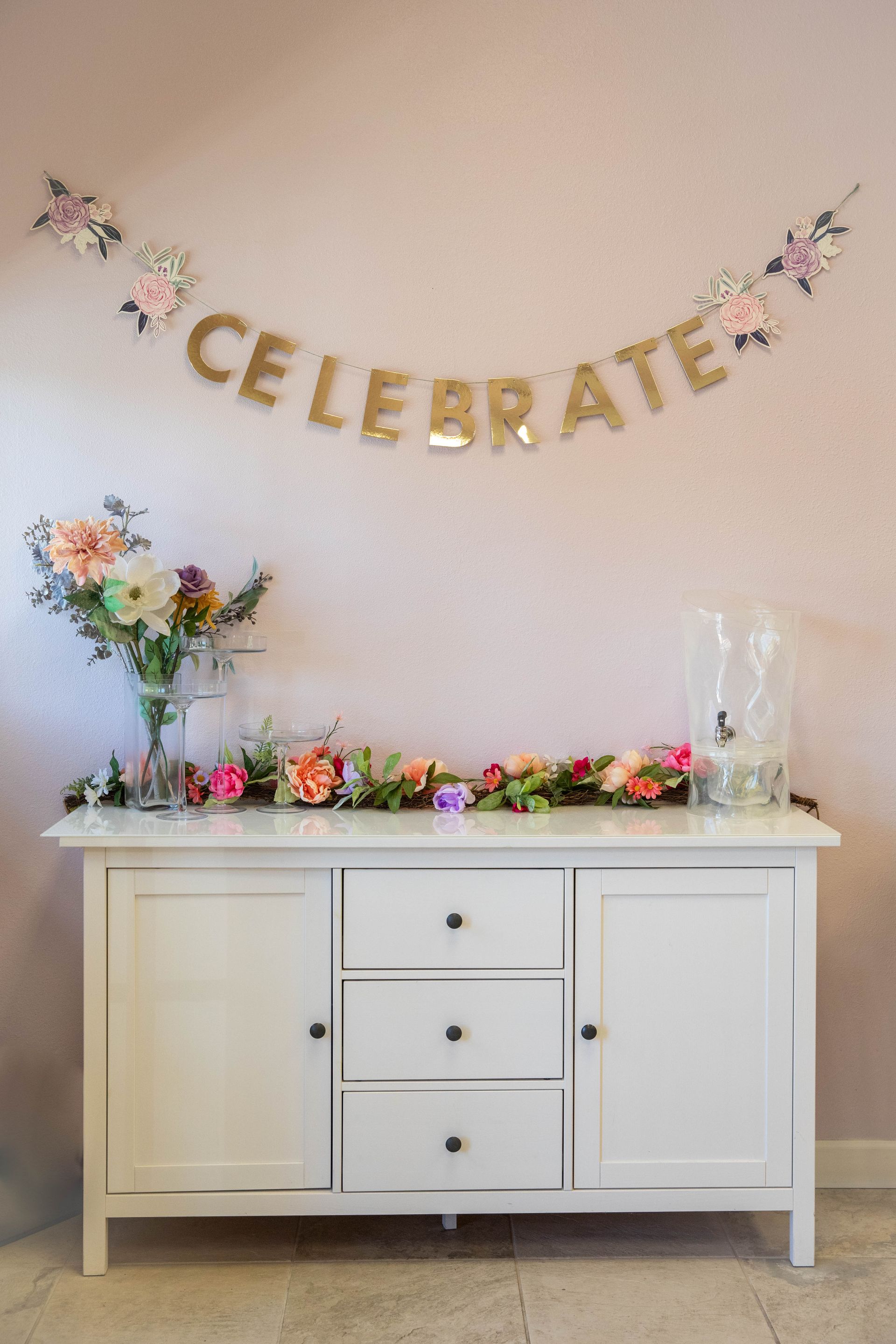 Decorated buffet table beneath a gold “CELEBRATE” banner with flowers and glass vases