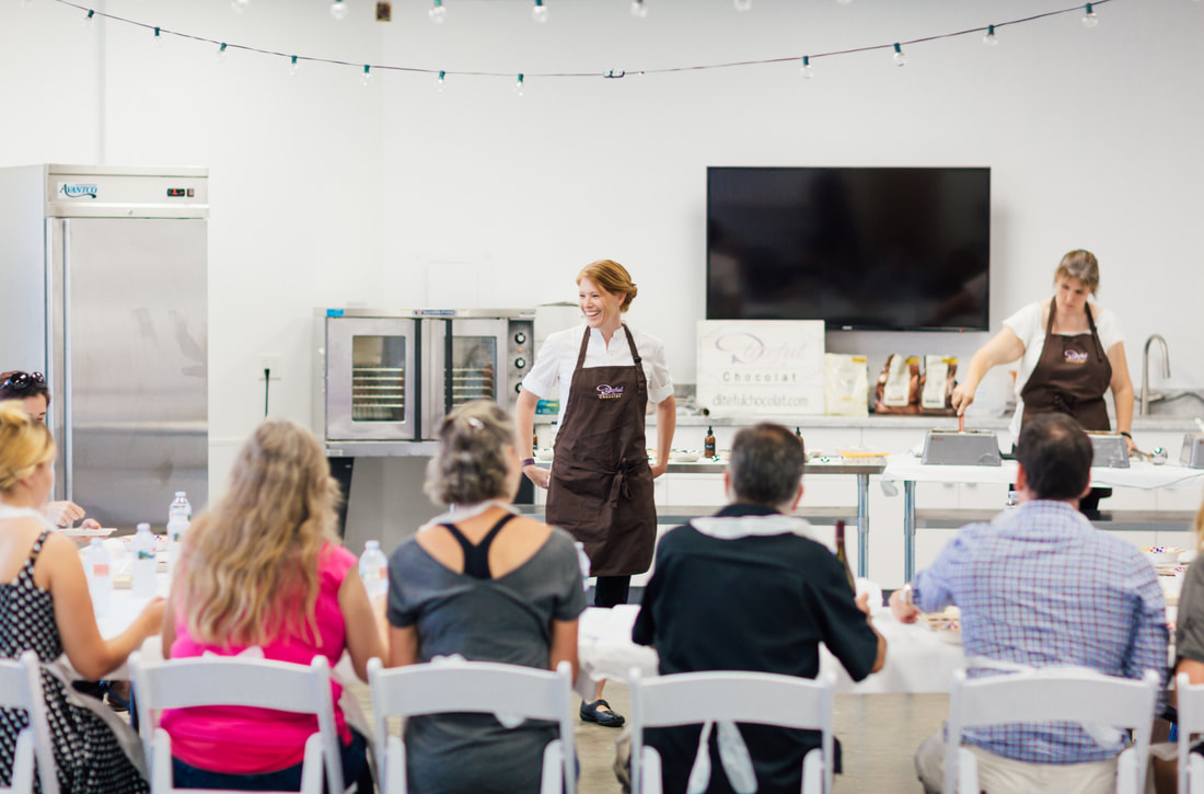 Cooking class with a presenter beside a large TV, facing seated attendees in a bright kitchen classroom.