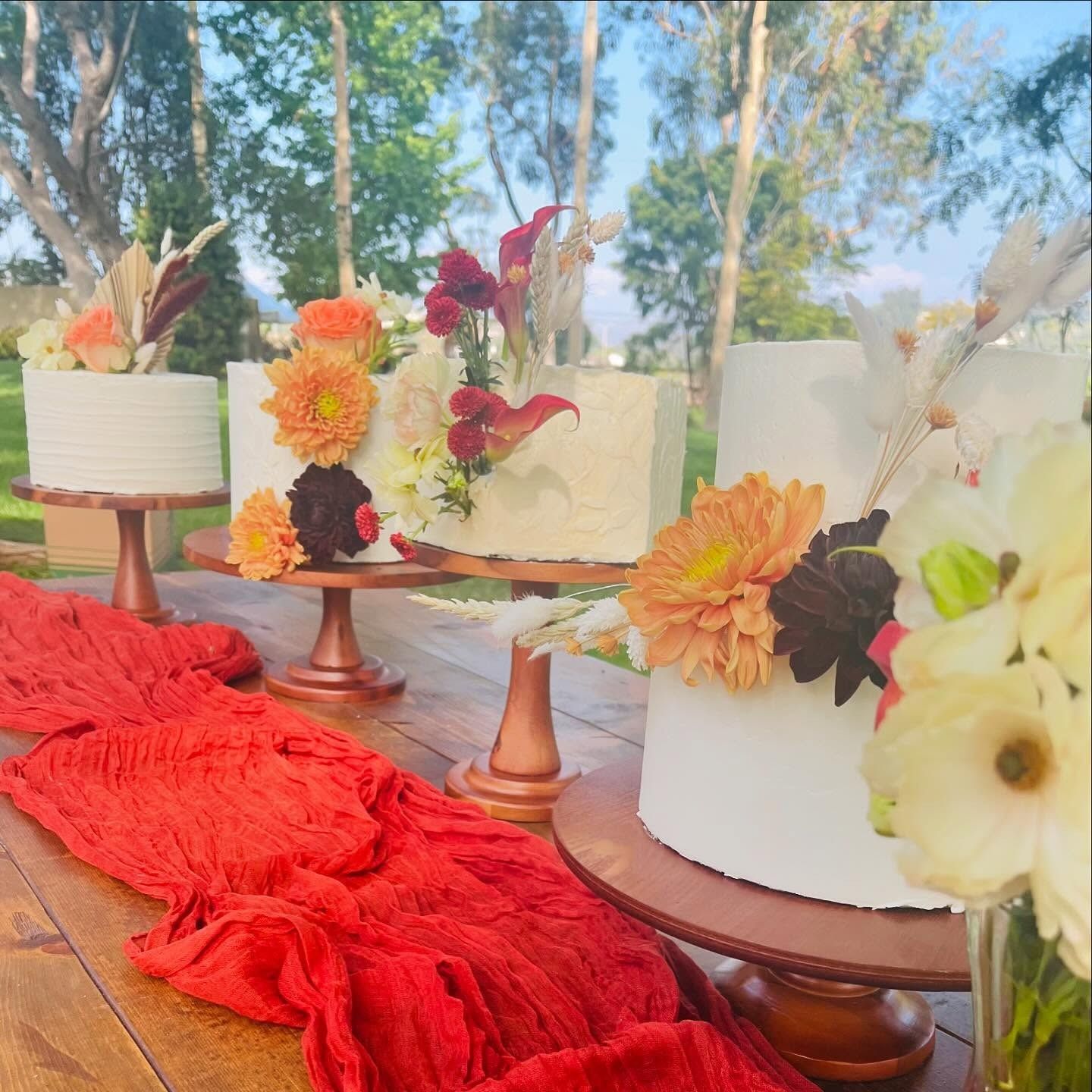 Floral-decorated white tiered cakes on wooden stands, arranged on a red tablecloth outdoors under trees.