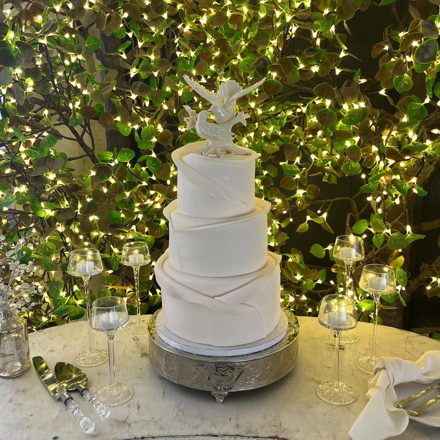 White tiered wedding cake with a bird topper on a silver stand, set on a decorated outdoor table under greenery