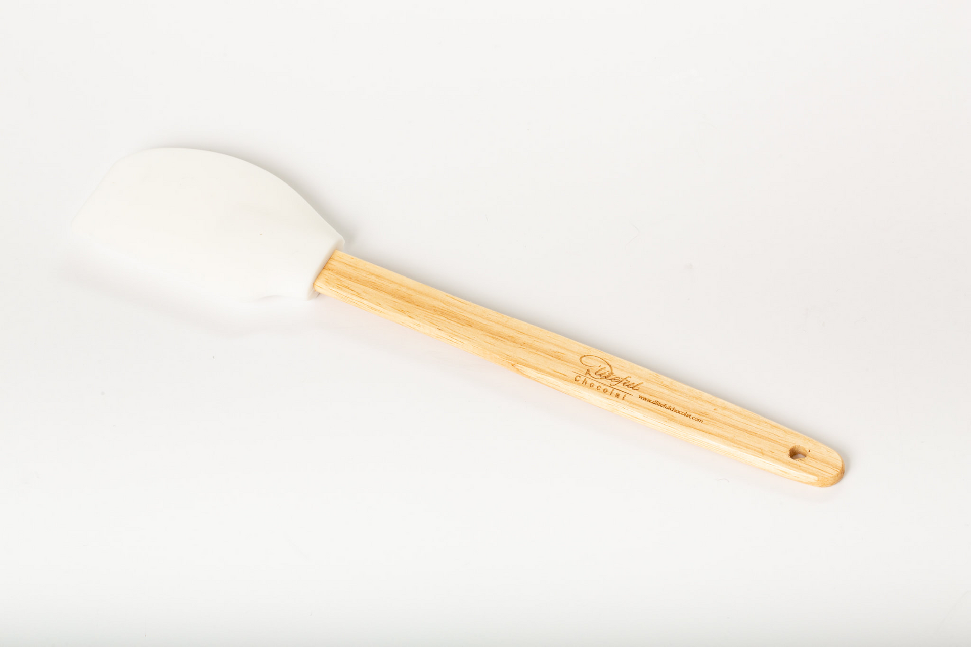 White silicone spatula with a wooden handle on a beige woven placemat, packaged in clear plastic.