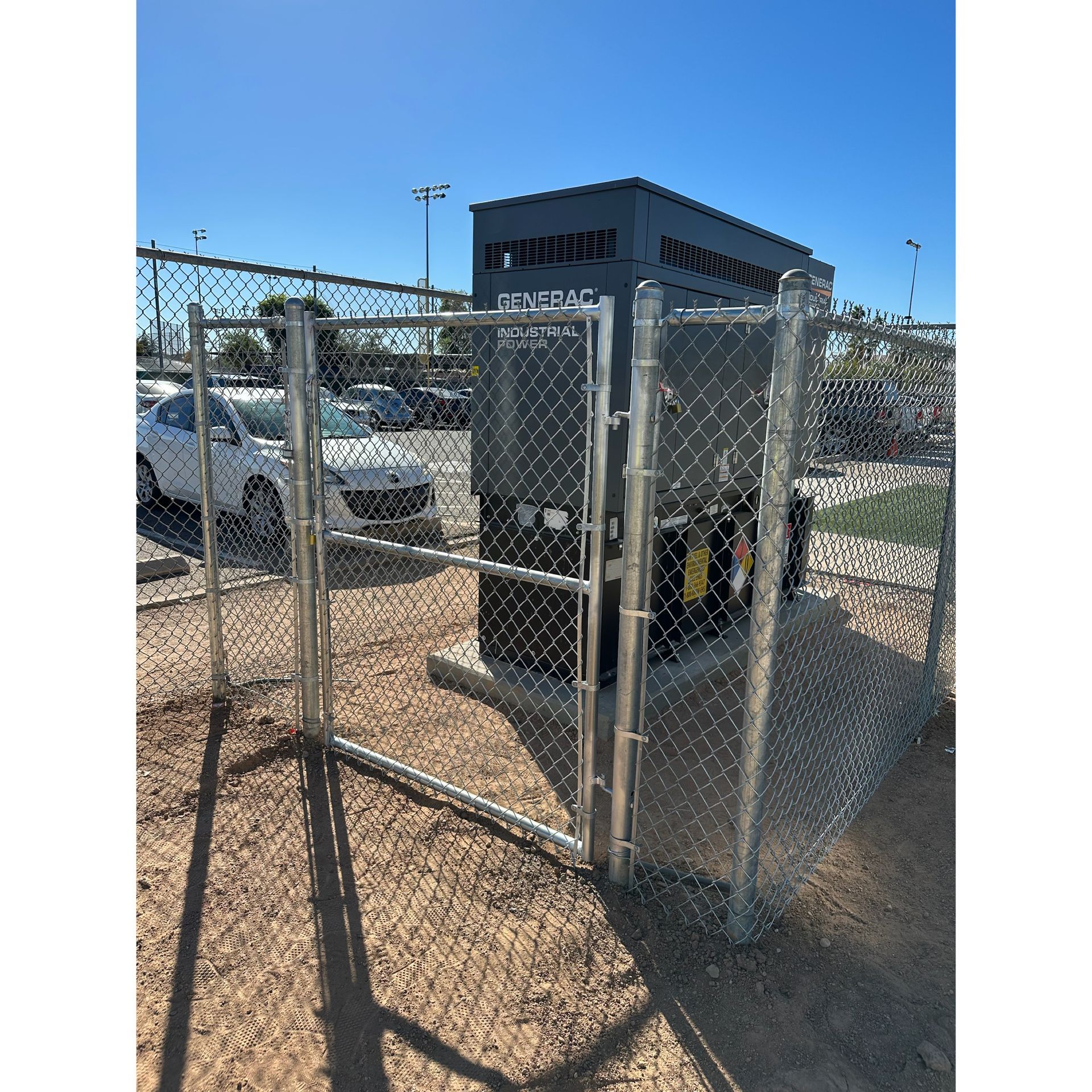 Chain-link fence enclosure with a black utility unit and open gate in a sunny parking lot