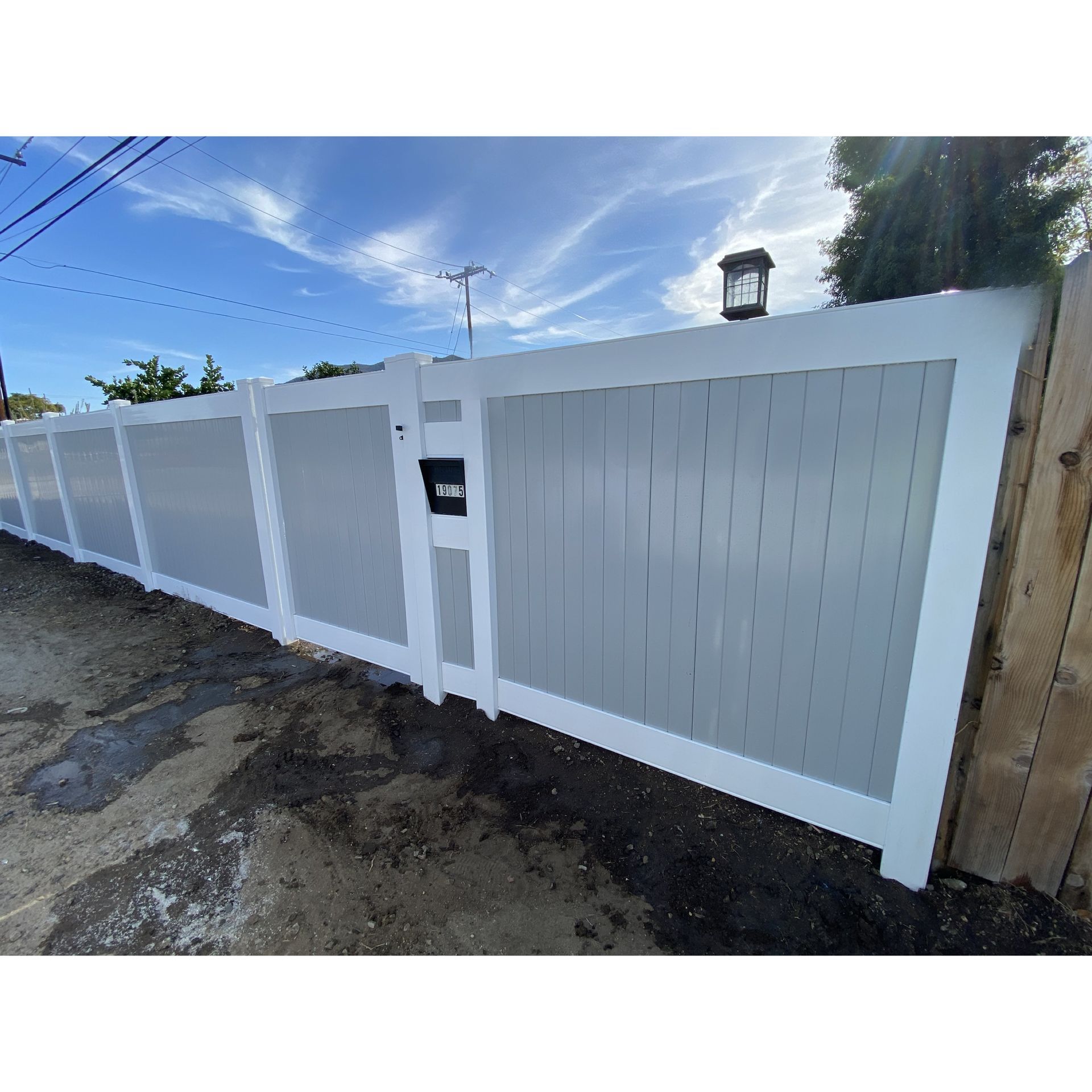 White fence and gate along a narrow street under a blue sky, with utility lines overhead.