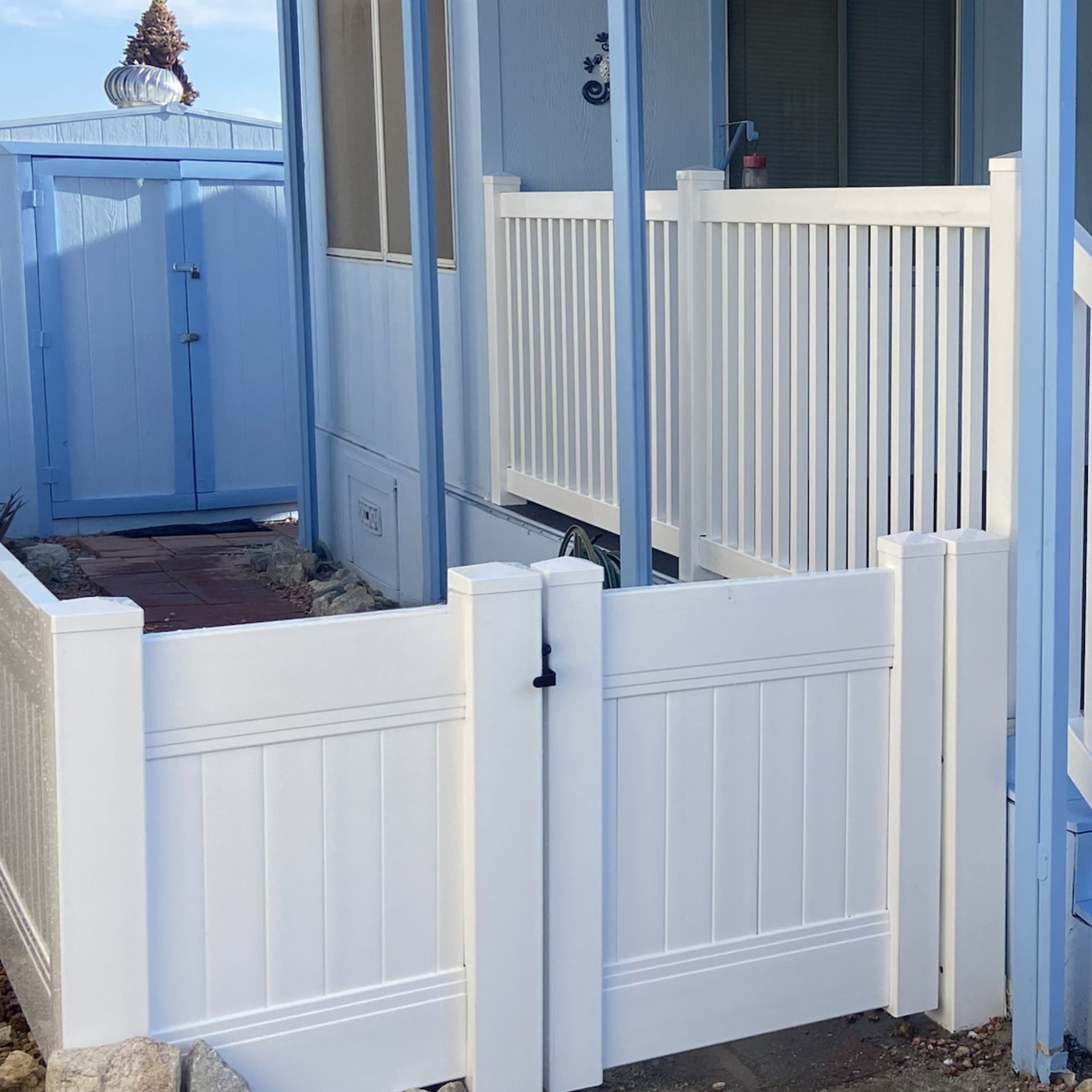 White vinyl gates and fence panels between blue buildings, with a narrow walkway and a potted plant on top.