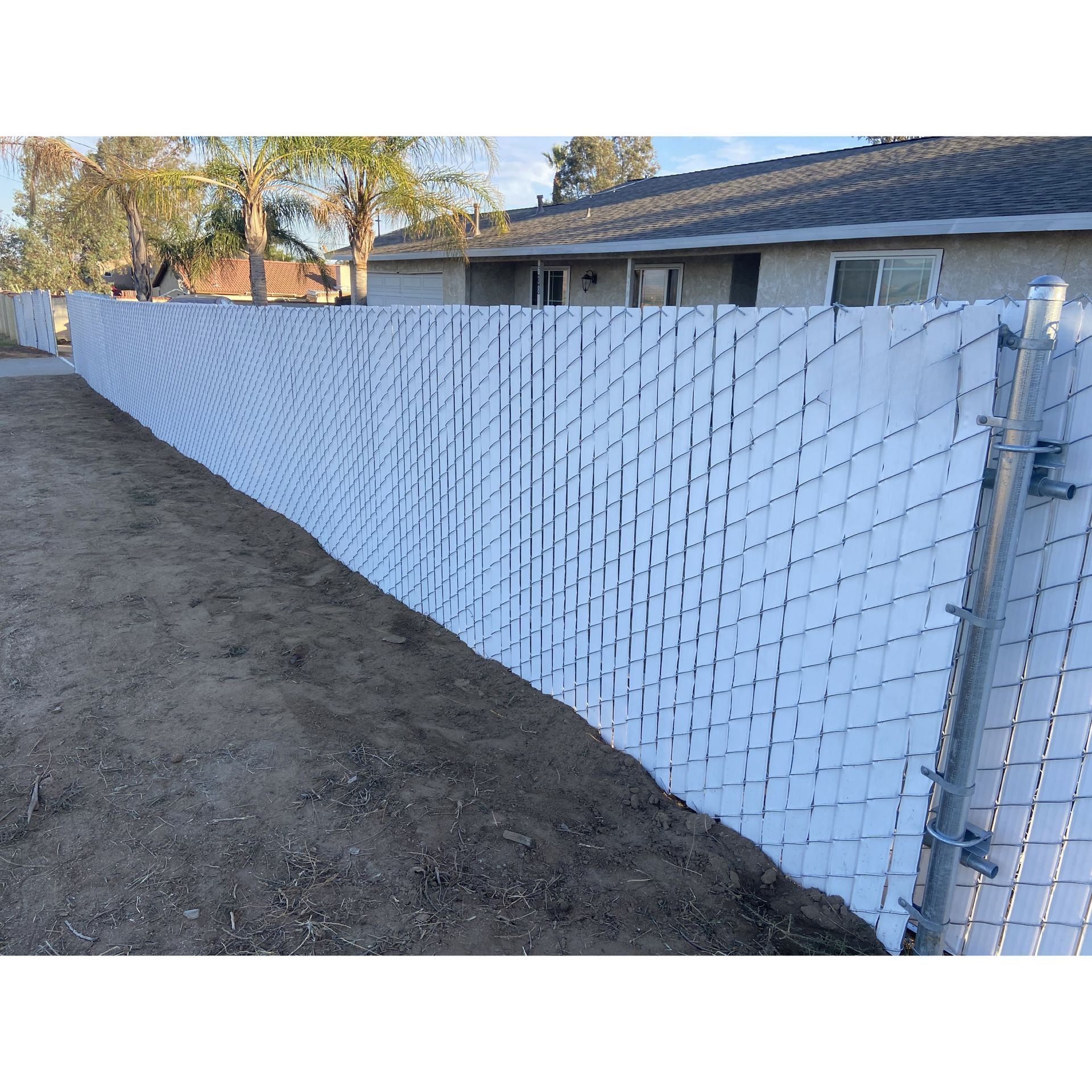 White fence along a dirt lot beside a single-story house under a clear sky