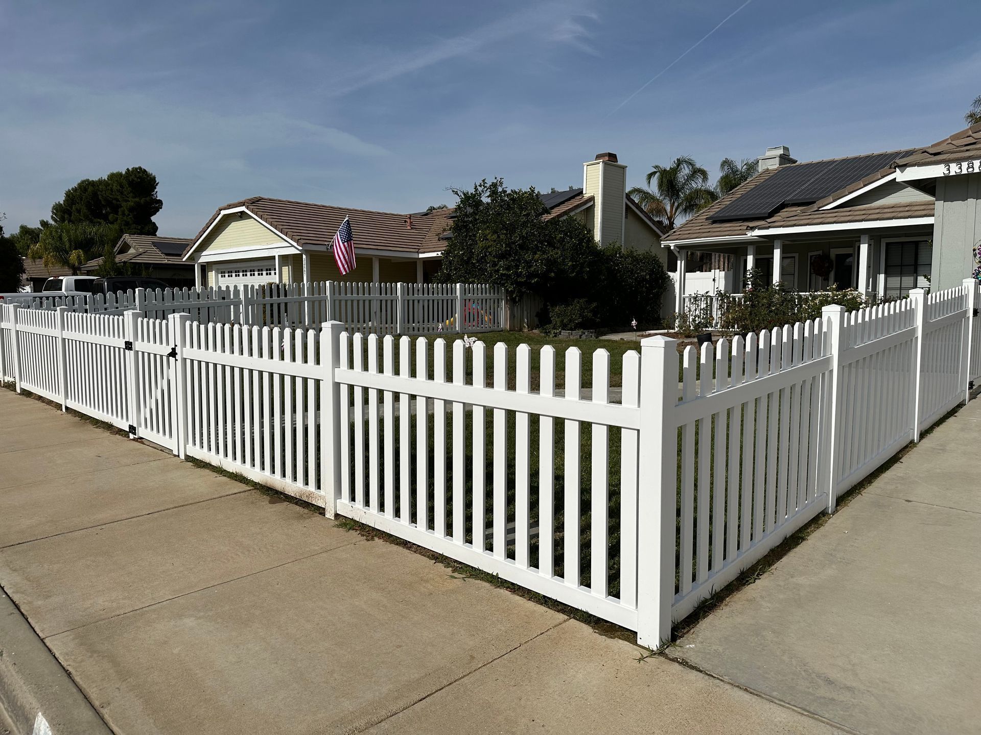 White picket fence enclosing a suburban corner lot with a house and driveway under a clear sky