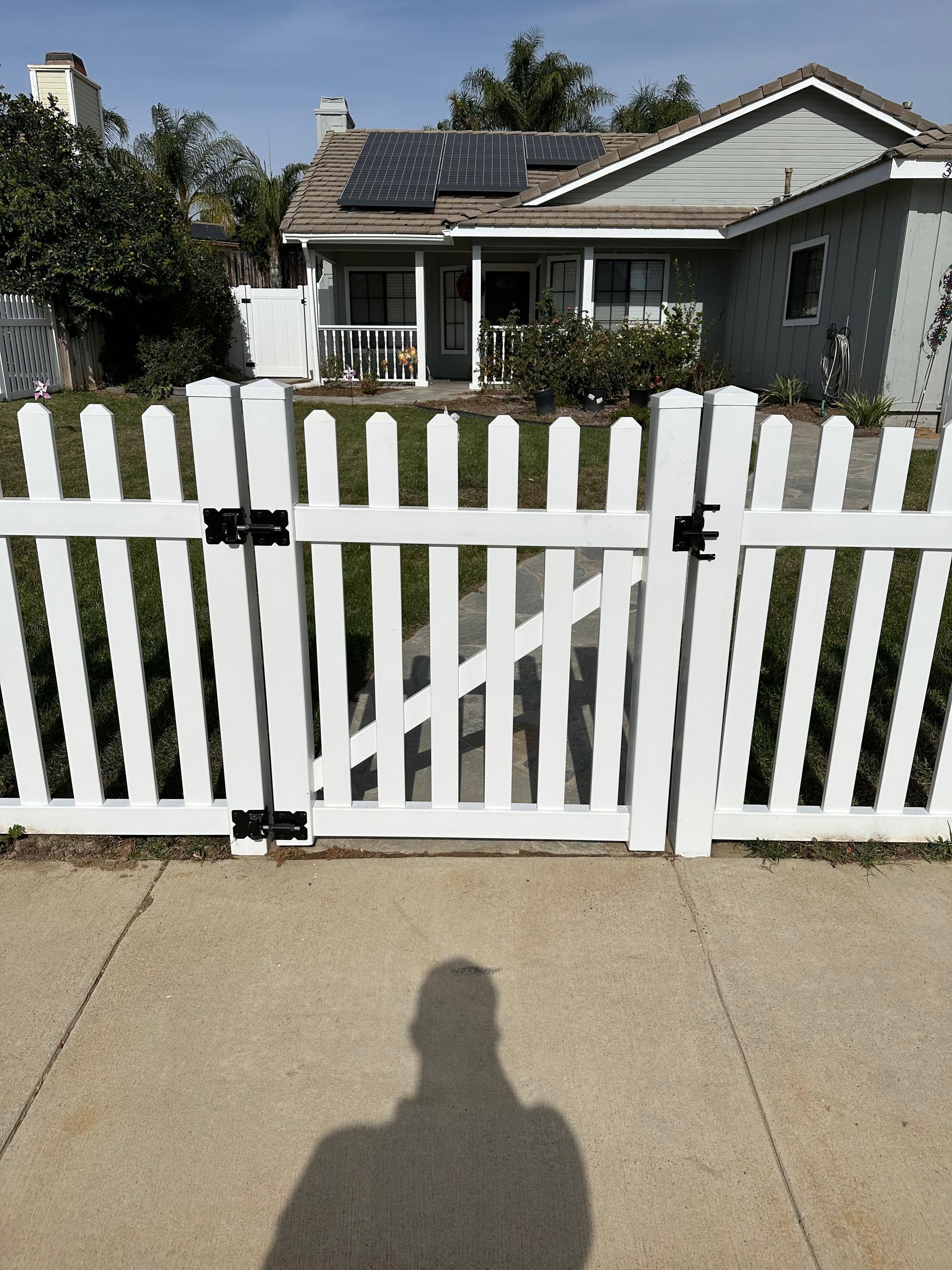 White picket fence in front of a house with solar panels, viewed from the sidewalk.