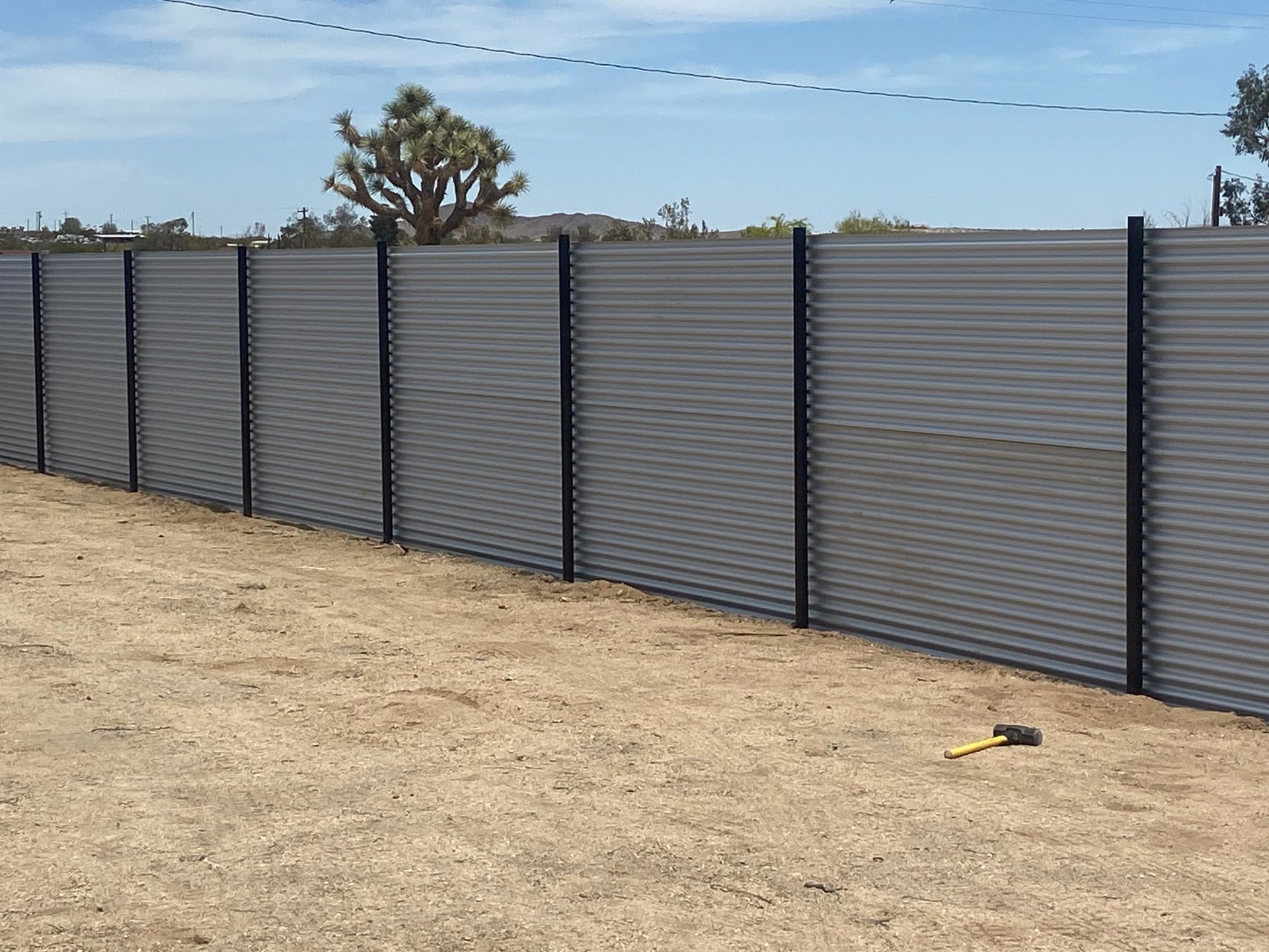 Gray corrugated metal fence along a dry dirt lot under a blue sky