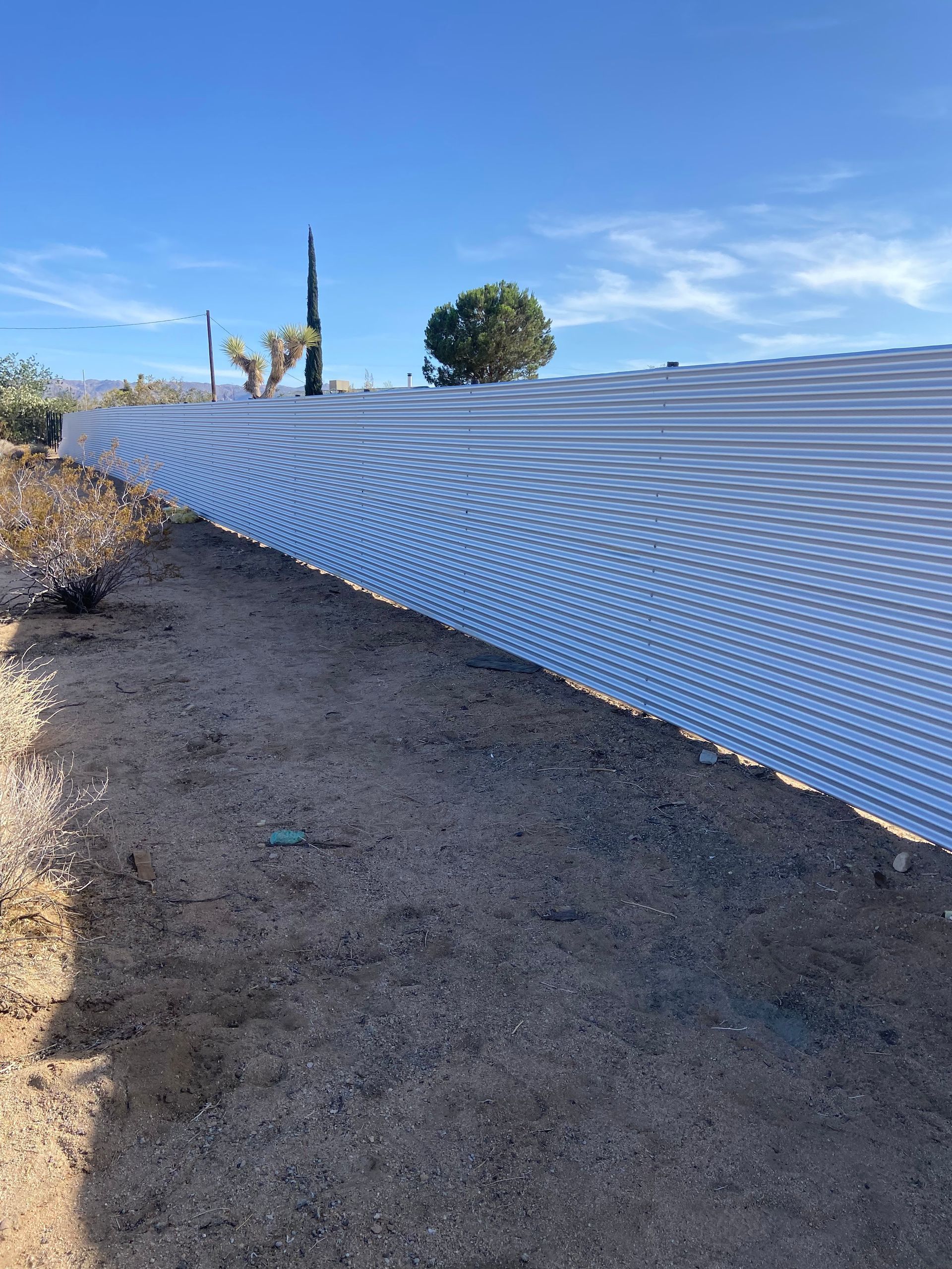 White corrugated fence along a dirt path under a blue sky, with desert plants in the background
