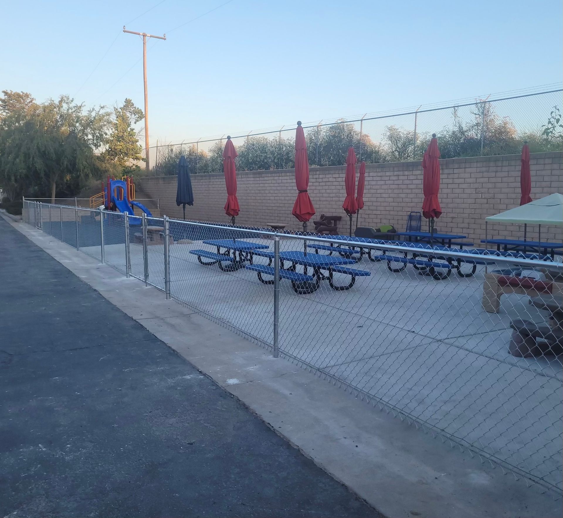 Outdoor patio with blue picnic tables and closed red umbrellas behind a chain-link fence