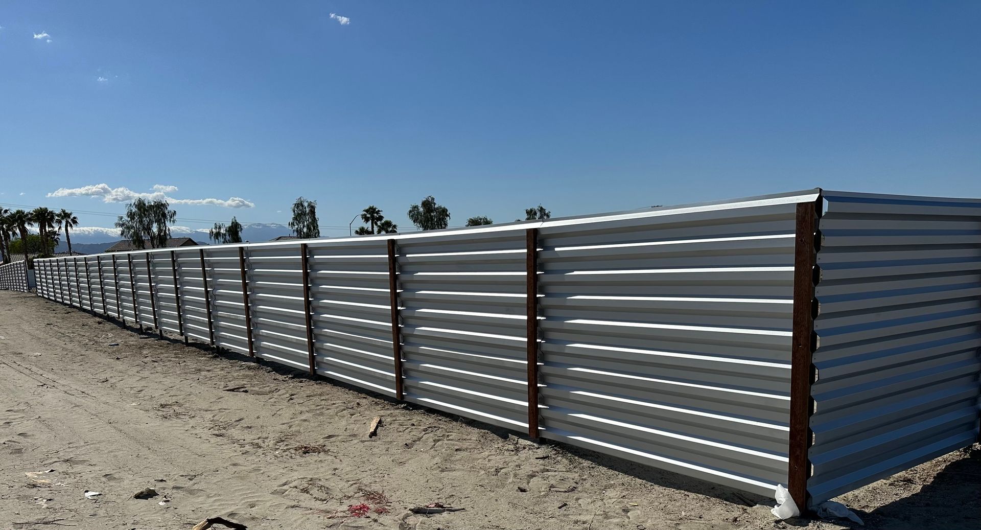 Long corrugated metal fence along a dirt road under a blue sky, with distant mountains and trees