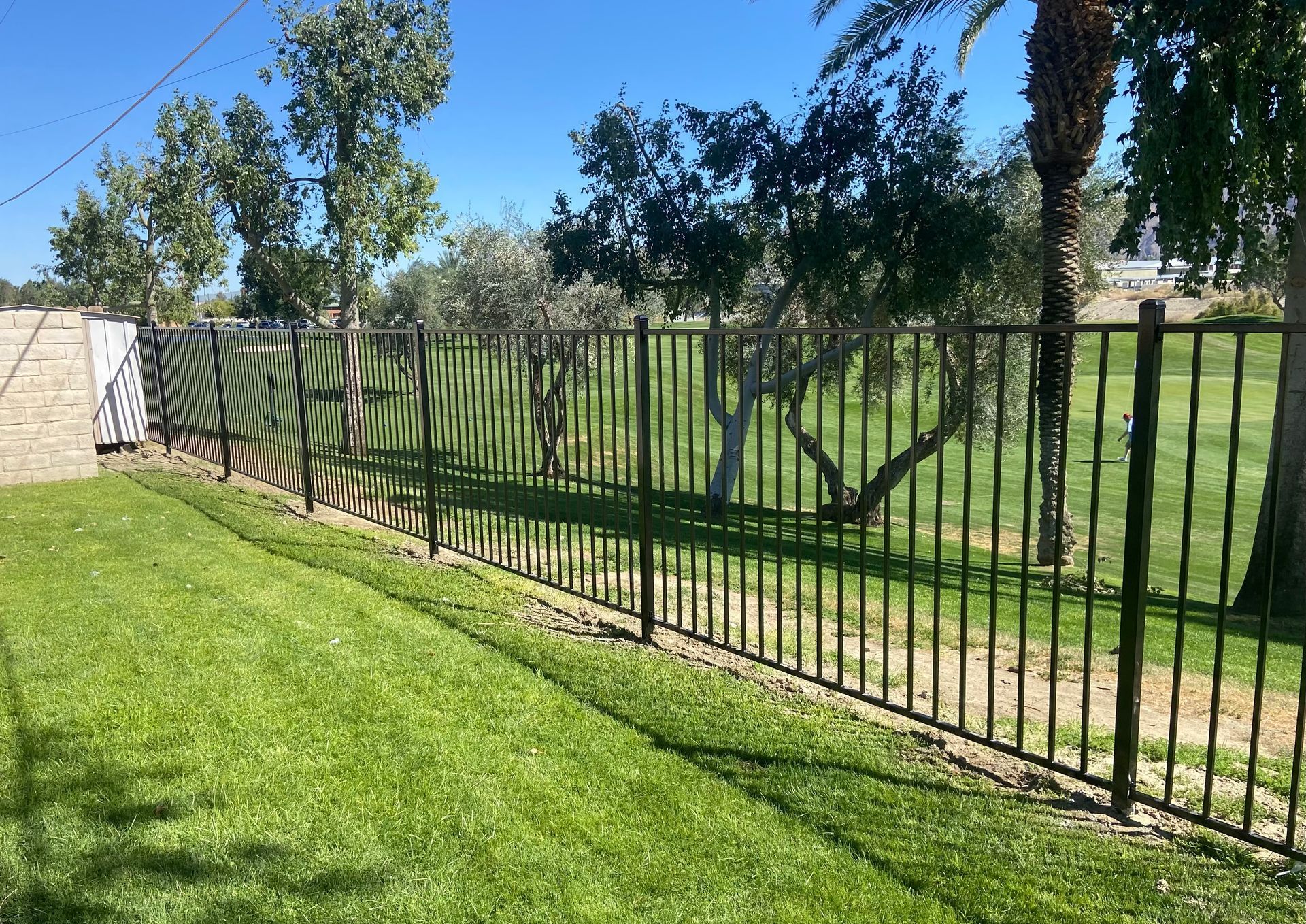 Black metal fence along a green lawn beside a stone wall under a sunny blue sky.