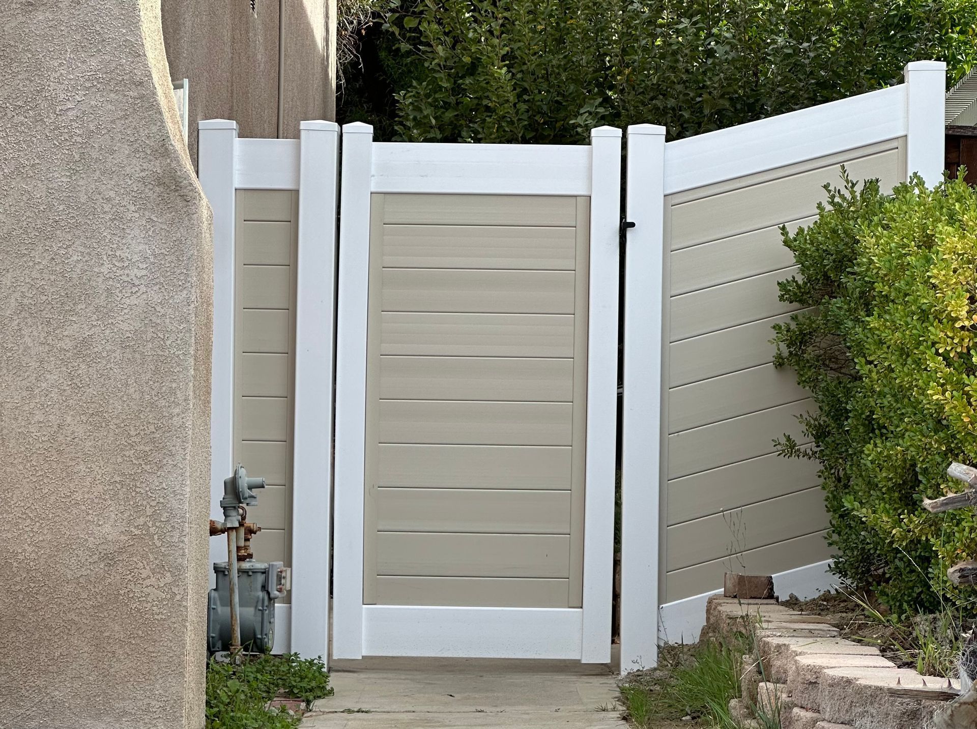 Beige privacy fence gate with white posts beside a stucco wall and shrubs