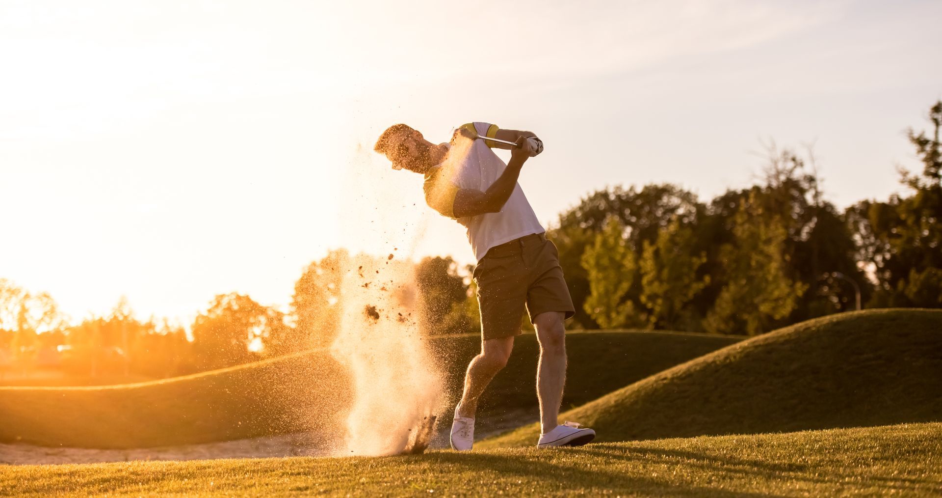 Golfer swings club, kicking up sand, on a sunny golf course.