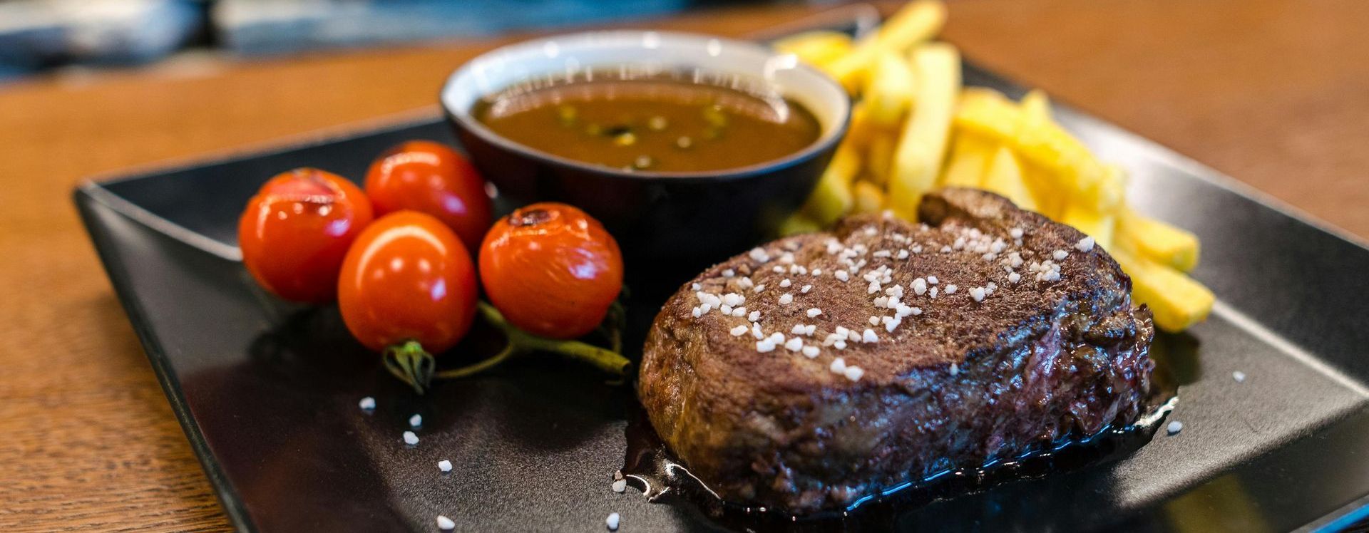 A black plate topped with a steak , french fries , tomatoes and sauce.