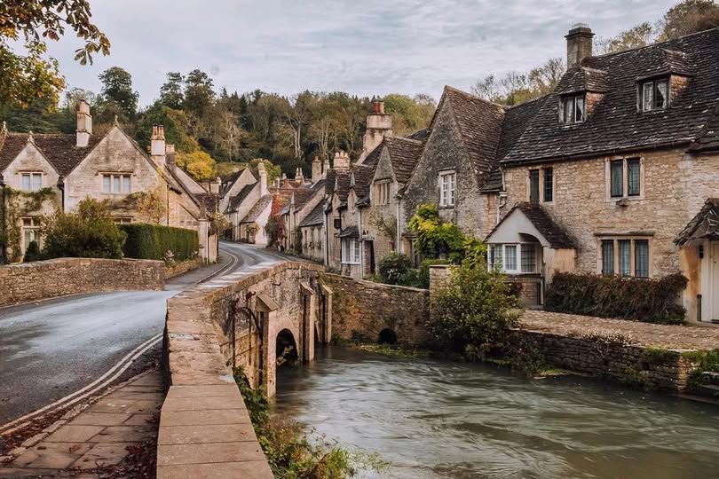 A road bridge crosses a stream with Cotswold stone houses lining the side of the water