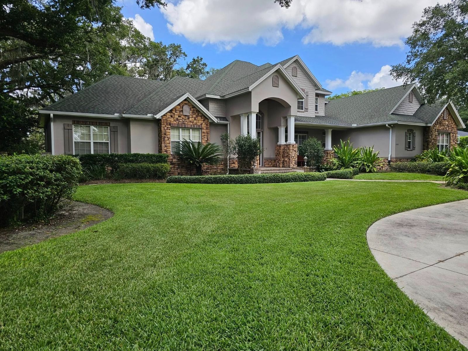 Large suburban house with gray roof, brick accents, and a manicured green lawn under a blue sky