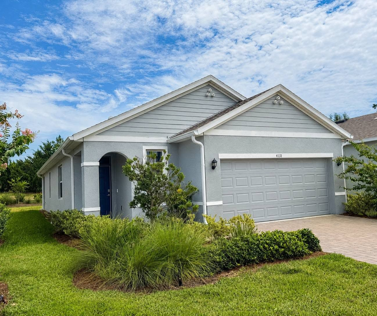 Single-story gray suburban house with blue front door, garage, and green lawn under a partly cloudy sky