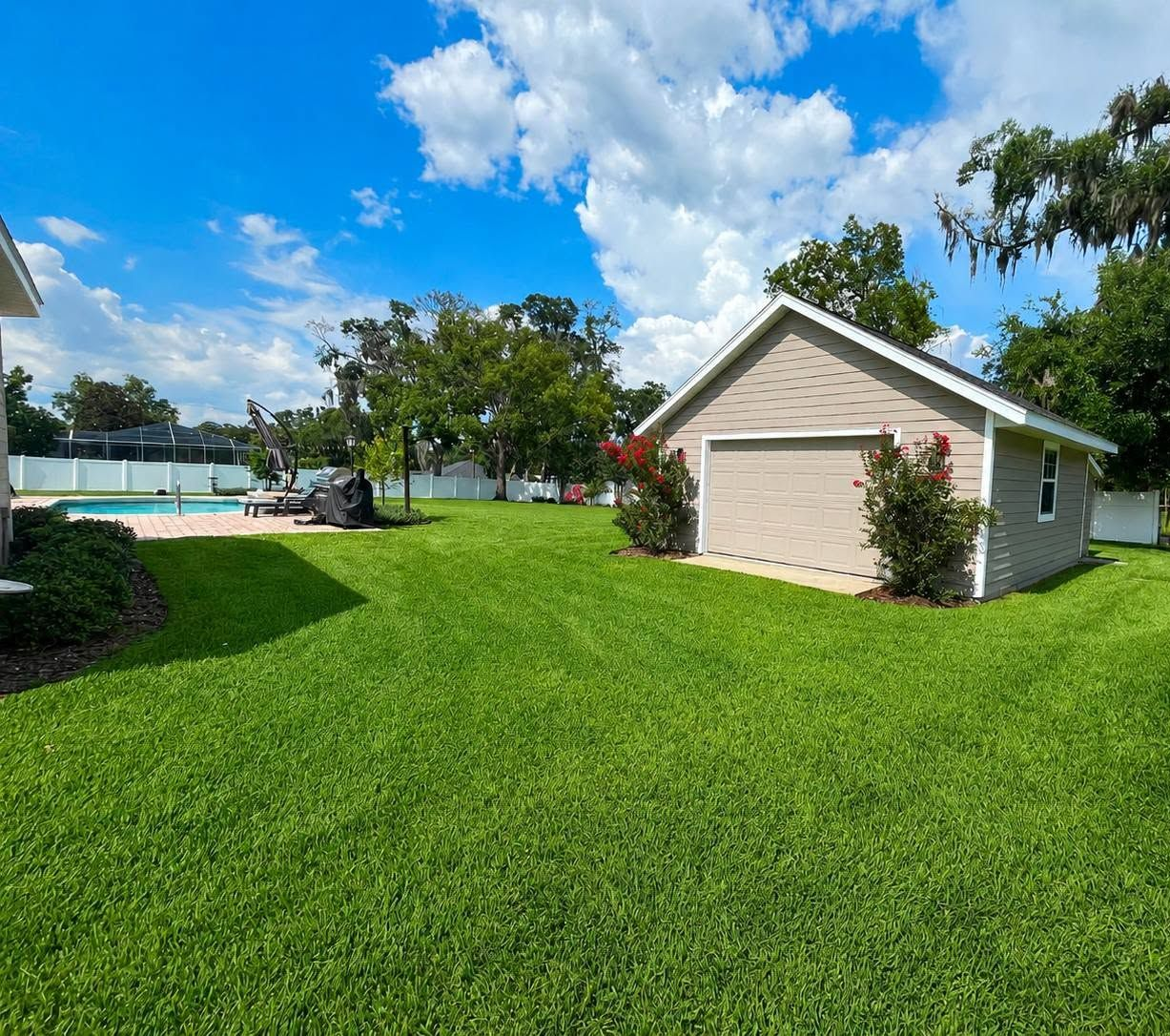 Sunny backyard with lush green lawn, a beige garage, and a swimming pool under a blue sky.