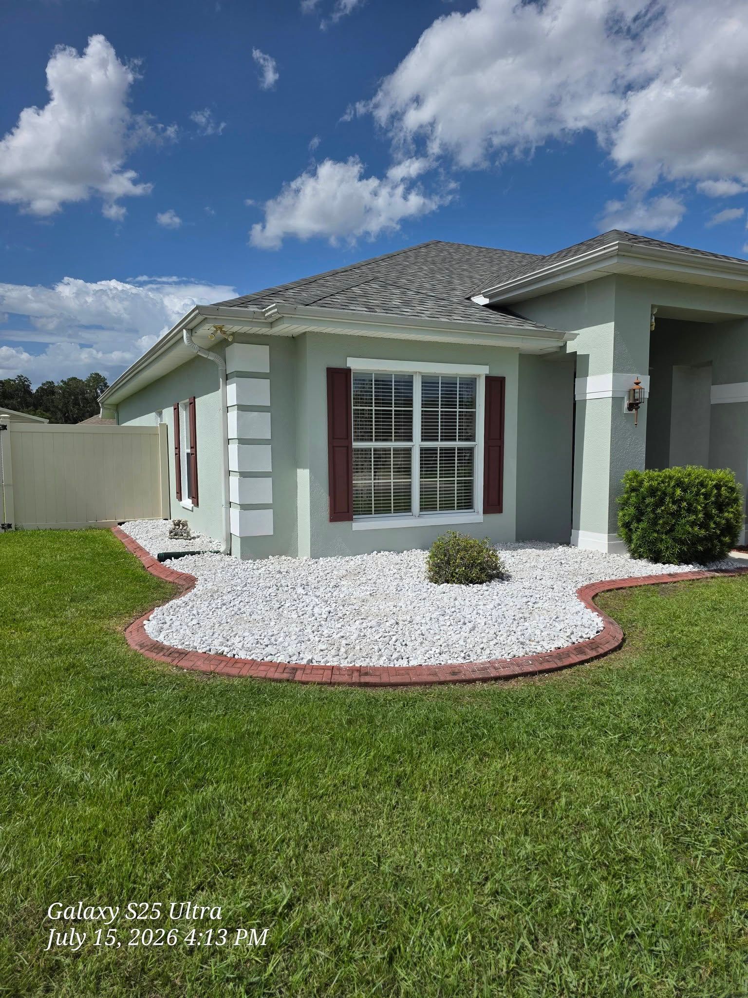 Single-story house with green trim, white stone landscaping, and a bright blue sky.