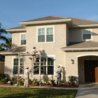 Two workers painting a beige two-story house exterior while one stands on a ladder